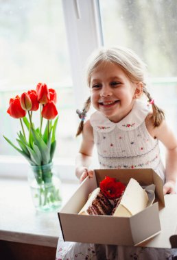 Dört yaşında güzel bir kız, bir odanın pencere eşiğinde oturur ve elinde çikolatalı pastayla bir kutu tutar. Camda bir buket kırmızı lale var. Beyaz işlemeli elbiseli sevimli sarışın.