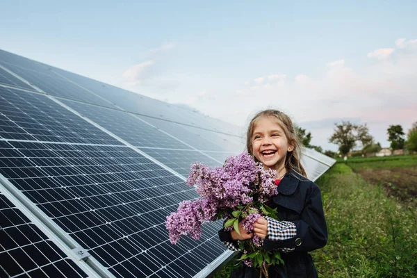 Alternatif enerjisi ve sürdürülebilir enerjisi olan bir çocuk. Çocuk çiçekleri güneş panellerinin arka planında tutuyor, fotovoltaik. Çevre dostluğu ve temiz enerji kavramı.