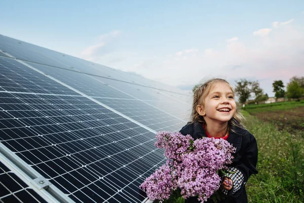 Alternatif enerjisi ve sürdürülebilir enerjisi olan bir çocuk. Çocuk çiçekleri güneş panellerinin arka planında tutuyor, fotovoltaik. Çevre dostluğu ve temiz enerji kavramı.