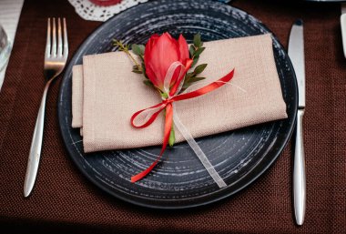 Bıçaklı boş siyah seramik tabak, kahverengi arka planda bir peçeteye çatal, kırmızı lale ile süslenmiş, yakın plan. Zarif bir sofra..