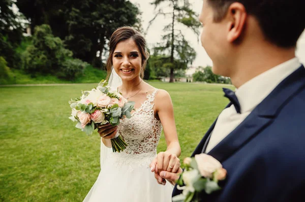 新郎手里拿着一束美丽的婚礼花束的笑容满面的新娘的画像 时尚的新婚夫妇在公园里散步 — 图库照片