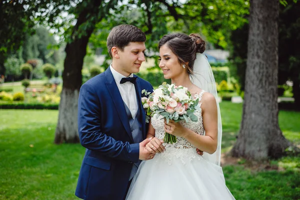 青いタキシードを着た新郎が白いドレスに身を包んだ花嫁を抱きかかえている 花嫁の手にピンクと白の牡丹の美しい結婚式の花束 — ストック写真