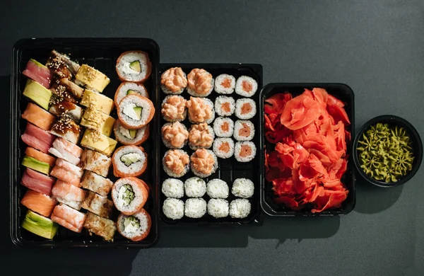 Japon gıda kavramı. Catering, suşi plaka veya plakalı çeşitli türdeki ayarlayın. Yemek çubukları, zencefil, soya sosu, wasabi. Plastik teslim kutusundaki taş siyah arka plan üzerine yerleştirilen.