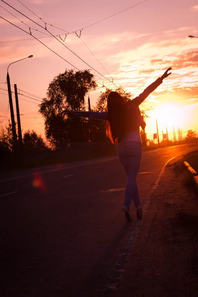 Jovem mulher em pé na estrada e olhando para um nascer do sol com as mãos levantadas. Liberdade de conceito . — Fotografia de Stock