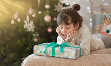 Komik, koyu saçlı bir kız hediyelerle yatakta oturuyor, Noel ağacının yanında. Yastığın üzerine yeni yılınız kutlu olsun! Bir yazıt için yer.