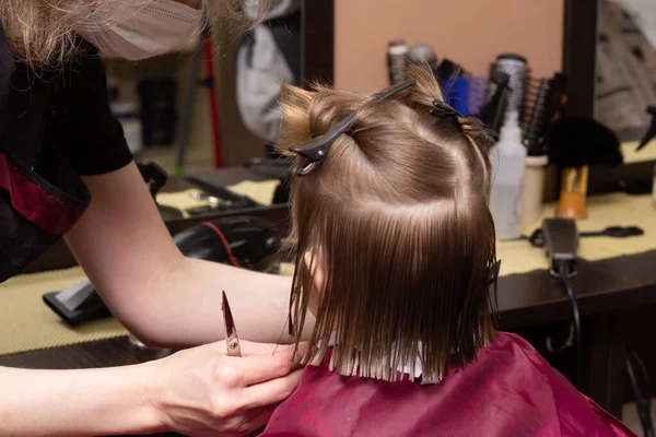 A little blonde girl 3 years old gets a bob haircut in a hairdresser. Baby hair care. Close-up.