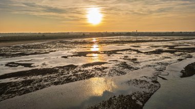 Fransa 'nın Normandiya kıyısındaki alçak gelgit sırasında su ve çamurlu düzlüklere yansıyan altın gün batımı. Sıcak tonlu huzurlu doğal manzara..