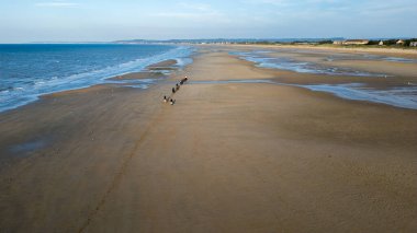 Dalgalar çekildiğinde kumlu bir sahil boyunca hareket eden at binicilerinin panoramik görüntüsü. Açık alan güzel bir Normandiya yaratırken, Atlantik dalgaları kıyıları silip süpürüyor. 