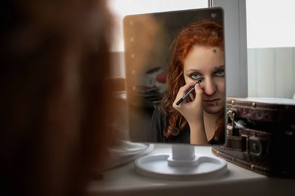 Aynaya bakarak makyaj yapan genç bir kadın. Kadın göz kalemi kullanıyor.