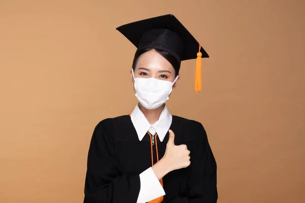 魅力的なアジアの女性はキャップとガウンを卒業し 医療マスク笑顔を身に着けているので 誇りと幸福のためにウイルスアウトブレーキとCovid 19ウイルスインフルエンザパンデミックベージュ背景 卒業コンセプトの新しい通常 — ストック写真