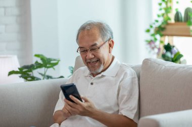 Evde cep telefonu ve sosyal medya tebessümü kullanarak koltukta oturan yaşlı mutlu Asyalı adam, ev konseptinde kıdemli yaşam tarzı.