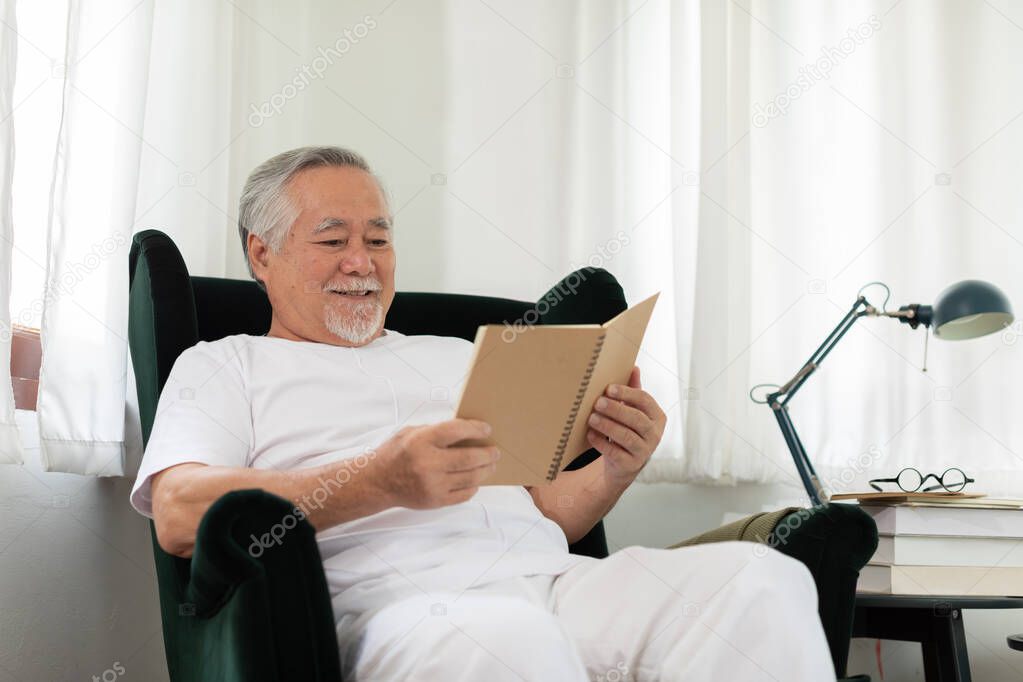 Asian Elderly man with white hairs sitting on sofa reading book ralax and cozy at home