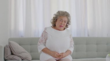 Yaşlı, Asya 'lı, beyaz saçlı yaşlı bir kadının evdeki kanepede mide ağrısı ve uti veya idrar kesesi ağrısı ve hastalığı vardır.