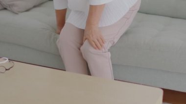 Beyaz saçlı yaşlı Asya 'lı yaşlı bir kadın diz ağrısı ve diz iltihabı, evinde kanepede gut, Yaşlı Bakım Konsepti var.