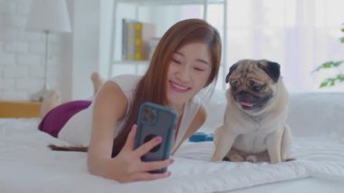 Güzel Asyalı genç bir kadın, şirin köpek yavrusuyla rahat bir yatak odasında fotoğraf çektirirken, tatilde evde dinlenmek için köpekle oynayan mutlu bir kız.