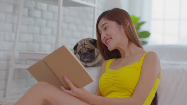 美しいアジアの若い女性読書本で犬で居心地の良いベッドルームリラックスして自宅で快適な レクリエーションのかわいい犬のパグ品種で休日一緒に — ストック動画