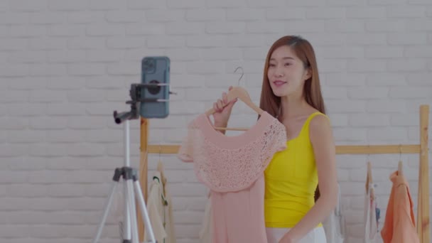 アジアの若い女性の起業家は オンラインプラットフォームやアプリケーションで顧客に彼女のドレスや服を販売するためのビデオをストリーミングライブ インフルエンサーかわいい女の子のレビューと自宅で布を提示 — ストック動画