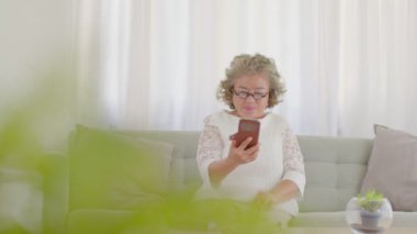 Kanepede oturan ve cep telefonu kullanan mutlu, yaşlı Asyalı kadın. VDO konferansı aile toplantısı ve evde gülümseme. Ev konseptinde kıdemli sağlık yaşam tarzı.