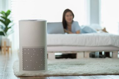Yatak odasında temiz toz ve temiz hava için hava temizleyici var. Bilgisayar laptopuyla çalışan ve arka planda rahatlayan bir kadın, nefes almak için temiz ve temiz havanın ve evde sağlığın iyi olması.