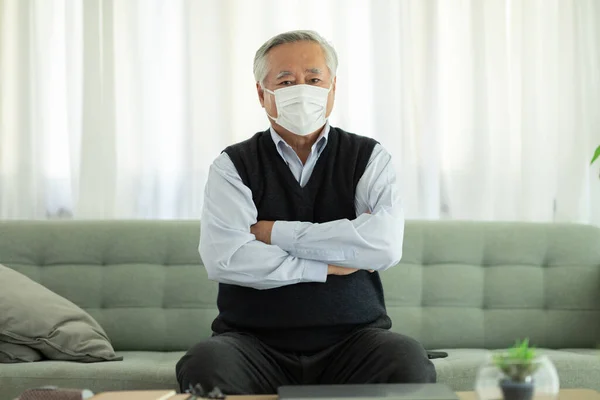 Happy Asian Elderly or Seniors man wearing medical mask to prevent coronavirus cross arms sitting on sofa in cozy living room at home,New normal lifestyle of confident older stay at home
