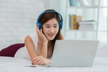 Mutluluk Asyalı genç bir kadın kulaklık takar yatağa uzanır müzik eşliğinde dinlenir, evde şarkı söyler rahat bir yatak odasında müzikle eğlenen genç bir kız