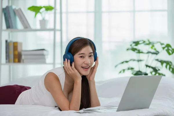 Happiness Asian young woman wear earphone lying on bed relax with music sing a song at home,Recreation young girl chilling with music in cozy bedroom