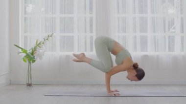 Asyalı bir kadının yeşil spor giysisiyle pencerelerin önünde yoga yaparken, yoga yaparken, yoga yaparken ya da Pincha Mayurasana pozunda, evde sağlıklı genç kadın solunumu ve meditasyon yogasının sakinliğinde.