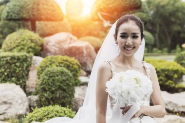 Güzel Asyalı gelin gelinlik giyip, gülümseyerek ve buket tutarak düğün gününde çok gururlu ve mutlu hissederek.