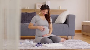 Mutlu Hamile Kadın elinde bez, beslenme şişesi, evde bebek ayakkabısı ile koca göbeğini okşayarak halının üzerinde oturuyor. Genç bir kadının gelecekten, annelikten ve gebelikten zevk almasının hamileliği.
