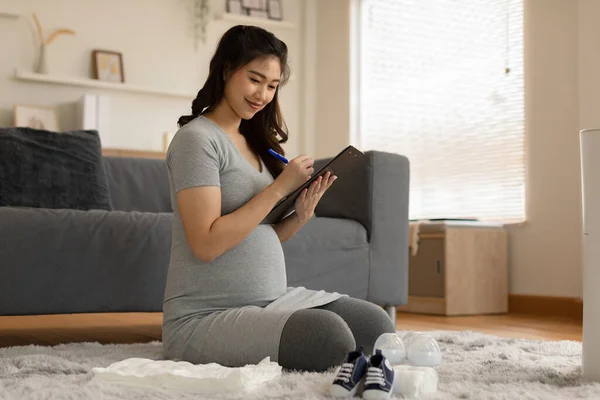 妊娠中の女性が自宅で赤ちゃんのために準備チェックリストを書いています妊娠中の女性は出産病院の準備ができて赤ちゃんのものを梱包します — ストック写真