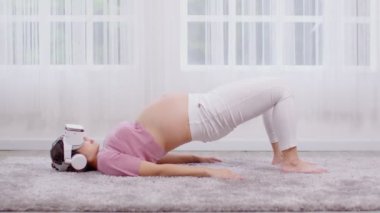 Mutlu Asyalı hamile kadın sanal gerçeklik gözlüklerini yoga dersi için kullanıyor. Yarı köprü, meditasyon gülümsemesine poz veriyor ve karnını evde rahatlatıyor. Hamilelik teknolojisi kavramı.