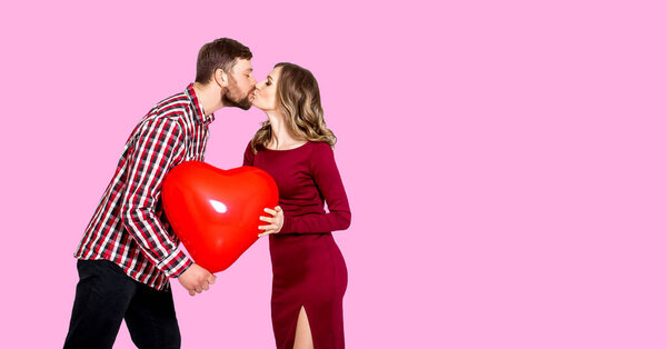 Girl and guy kiss on a pink background and hold in their hands a large inflatable red ball in the shape of a heart. Concept of the holiday - Valentine's Day, International Women's Day, Mother's Day