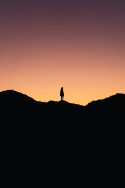 Dağın ortasında duran bir kişinin dikey fotoğrafı. Arka planda gün batımının renkleri var.
