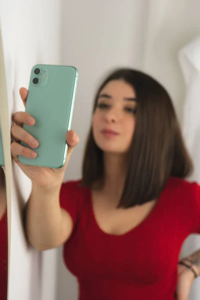 Kısa saçlı, kırmızı gömlekli genç bir esmer kız aynanın yanında cep telefonuyla selfie çekiyor.