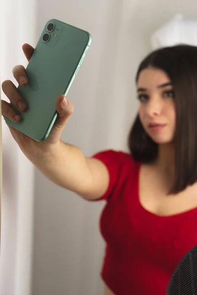 Kısa saçlı, kırmızı gömlekli genç bir esmer kız aynanın yanında cep telefonuyla selfie çekiyor.