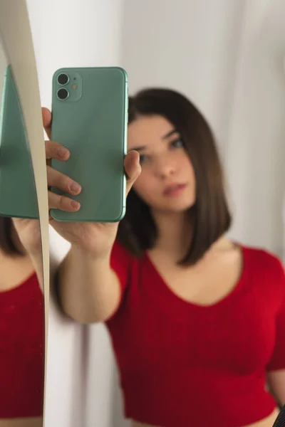 Kısa saçlı, kırmızı gömlekli genç bir esmer kız aynanın yanında cep telefonuyla selfie çekiyor.
