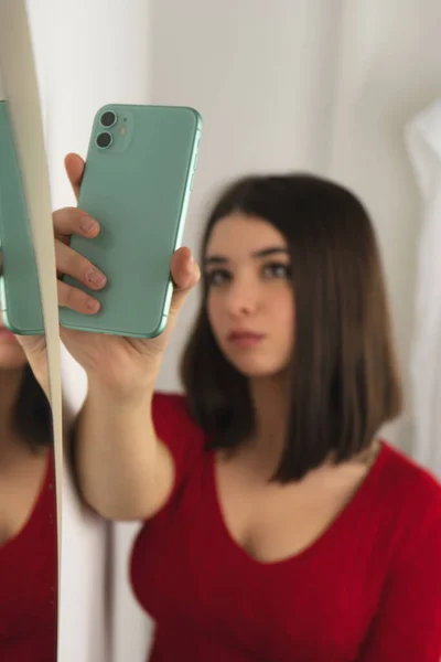 Kısa saçlı, kırmızı gömlekli genç bir esmer kız aynanın yanında cep telefonuyla selfie çekiyor.