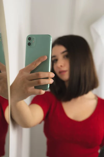 Kısa saçlı, kırmızı gömlekli genç bir esmer kız aynanın yanında cep telefonuyla selfie çekiyor.