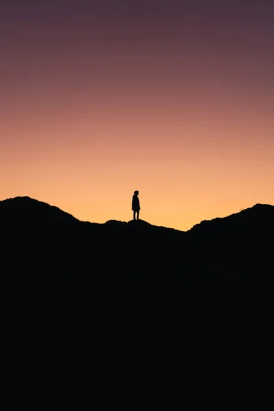 Dağın ortasında duran bir kişinin dikey fotoğrafı. Arka planda gün batımının renkleri var.