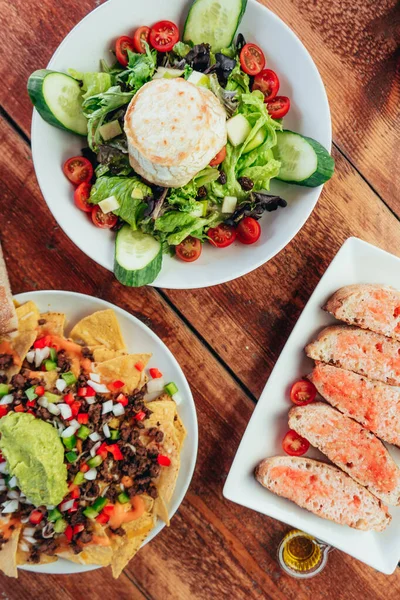 Keçi peynirli salatanın dikey fotoğrafı, guacamole soslu nacho ve ahşap bir masada domatesli et ve ekmek.