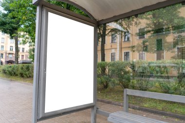 Bir afiş için dikey reklam panosu, yeşil bir ara sokakta, toplu taşıma durağında duruyor.