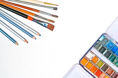 Beyaz kağıda, püsküllere, kalemlere, suluboya sanat malzemelerine düz resimler serilmiş.