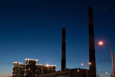 Gecenin geç saatlerinde mavi gökyüzüne karşı sigara içen evler ve endüstriyel borular.