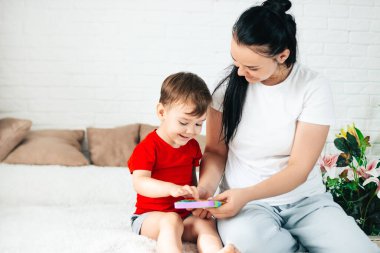 Bir anne ve 2 yaşındaki bir çocuk beyaz bir odada renkli bir pop-it oyuncağıyla oynuyorlar. Stres önleyici hassas bir oyuncak ya da tekrar kullanılabilir bir baloncuk paketi. 2021 trendi.