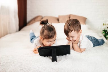 İki çocuk beyaz bir yatakta tablet bilgisayar kullanıyor. Beyaz bir odada tablet bilgisayarı olan bir kız ve bir oğlan.