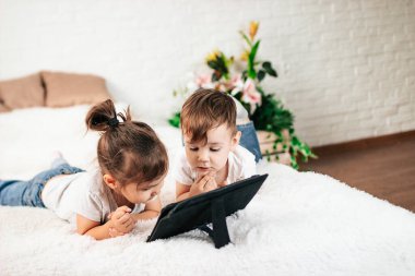 İki çocuk beyaz bir yatakta tablet bilgisayar kullanıyor. Beyaz bir odada tablet bilgisayarı olan bir kız ve bir oğlan..