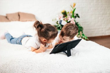 İki çocuk beyaz bir yatakta tablet bilgisayar kullanıyor. Beyaz bir odada tablet bilgisayarı olan bir kız ve bir oğlan..