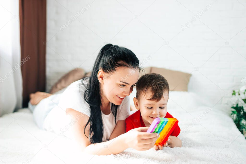 A mom and a 2-year-old boy play with a colorful fashion pop-it toy in a white room on the bed, an anti-stress sensitive toy or a reusable bubble wrap. The trend of 2021.
