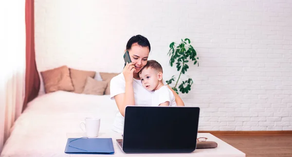a young black-haired mother with a small child works at home in a bright room with a laptop