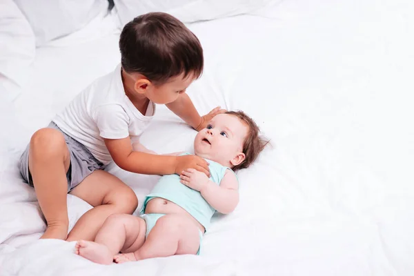 Симпатичный мальчик обнимает младшую сестру на белоснежной кровати — стоковое фото